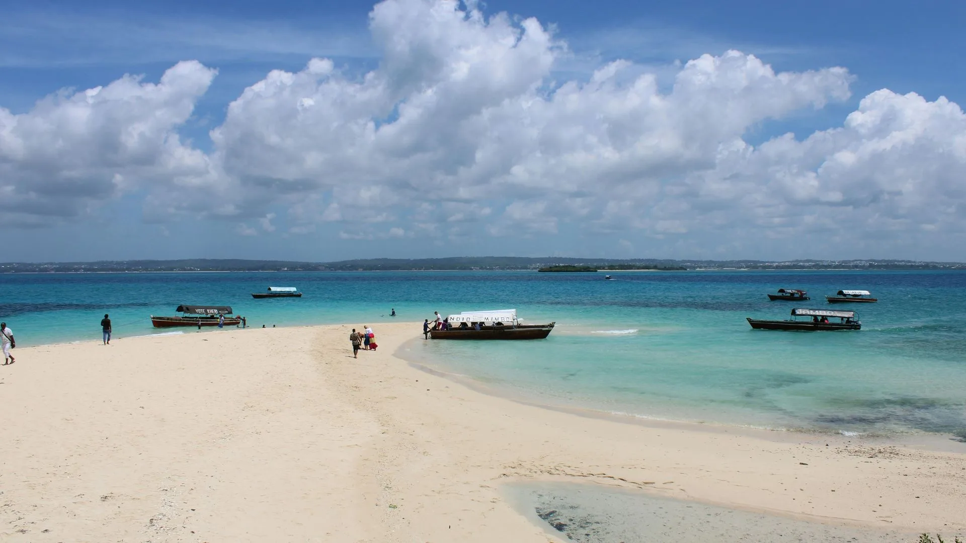 White-sand beach spit with turquoise water and traditional dhow and motorboats moored off a Zanzibar sandbank under scattered cumulus clouds