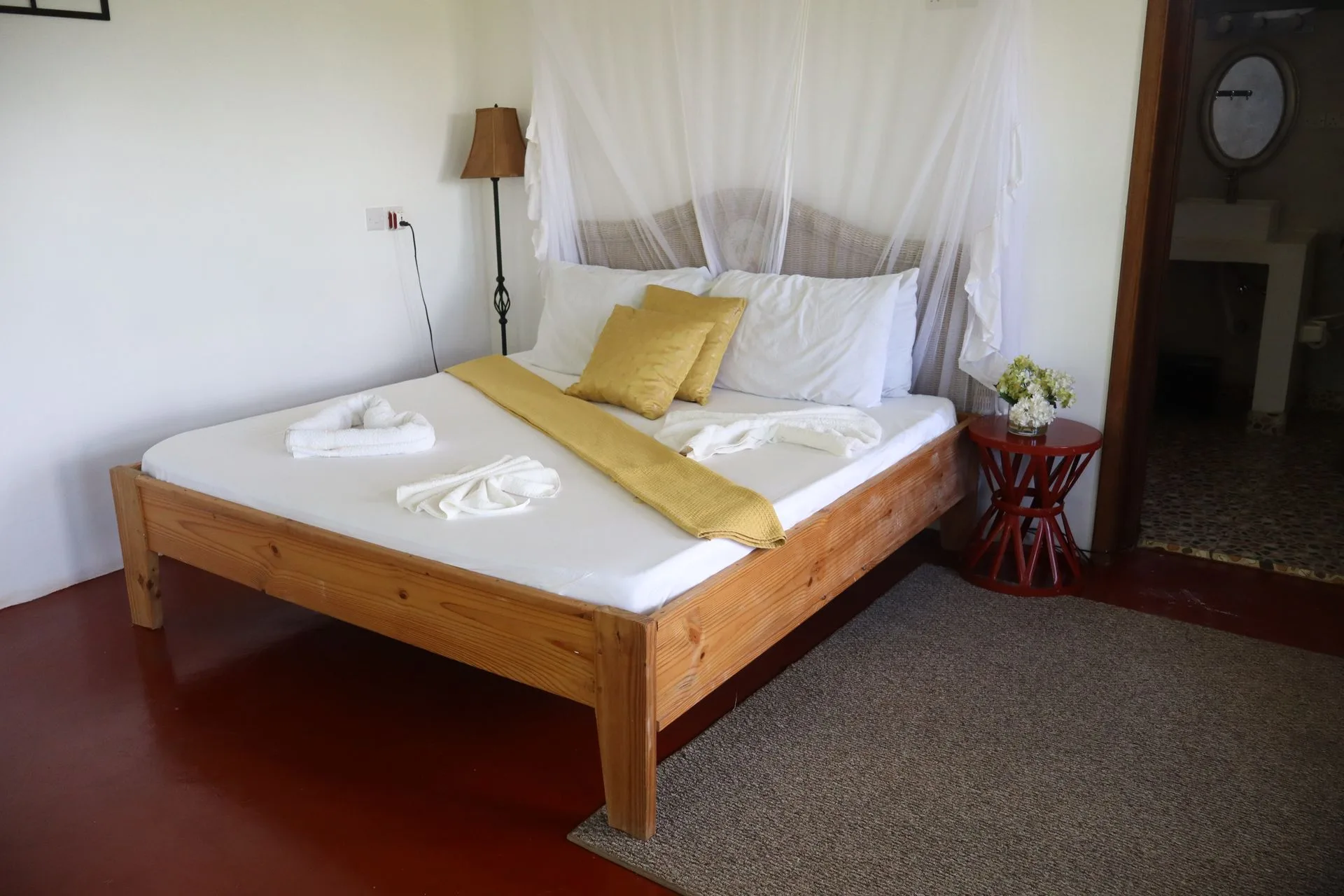 Sunlit bedroom at Hamia Zanzibar with a carved wooden canopy bed, flowing white mosquito net, polished red concrete floor and garden views through tall windows